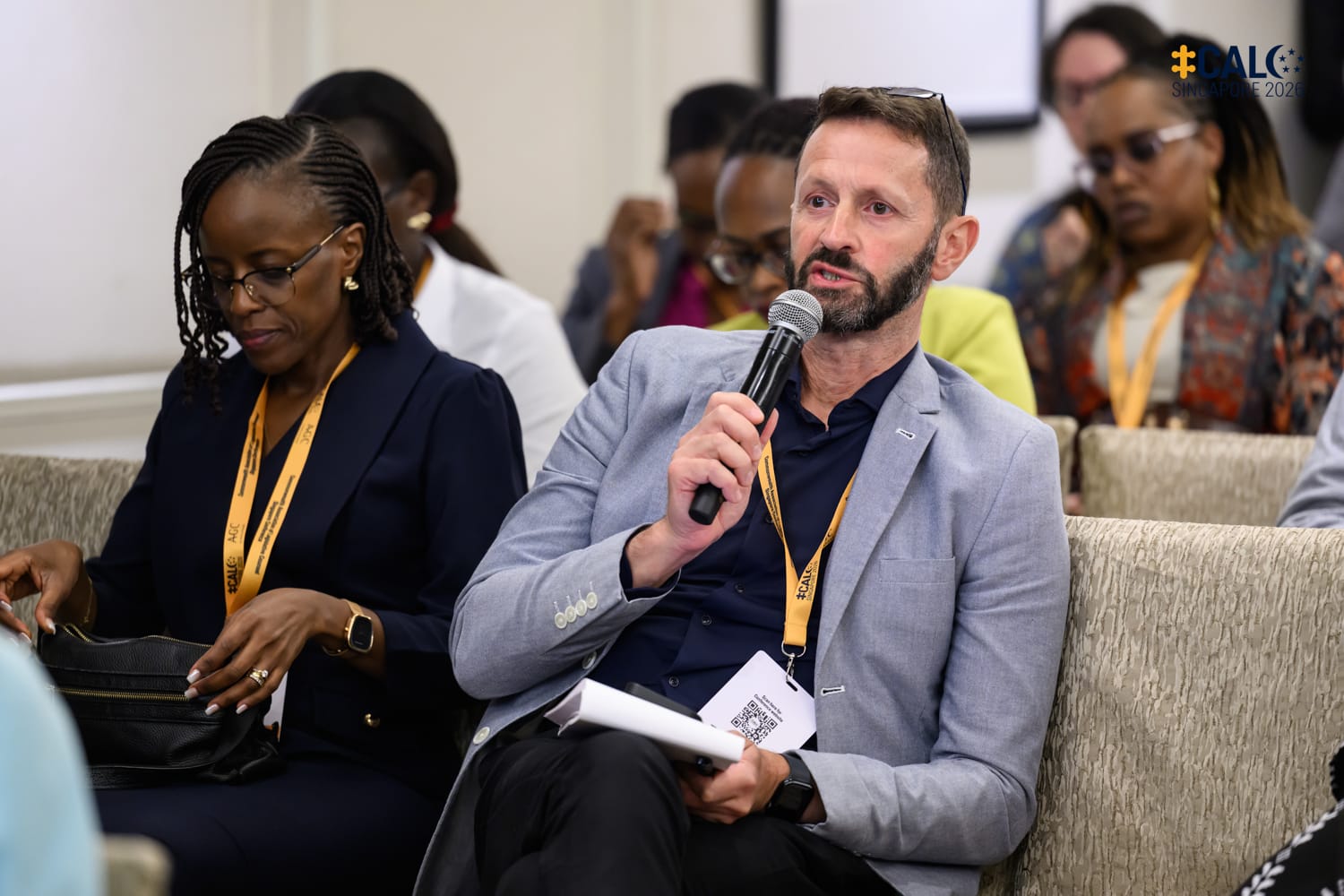 A delegate speaks into a handheld microphone during a Q&A session at the Conference, seated among other attendees in a breakout room at The Fullerton Hotel Singapore.